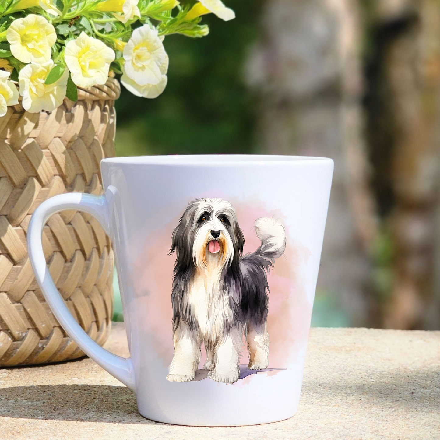 A white ceramic latte mug featuring a standing Bearded Collie in a pink mist background.  A basket containing yellow flowers sits to the left hand side.