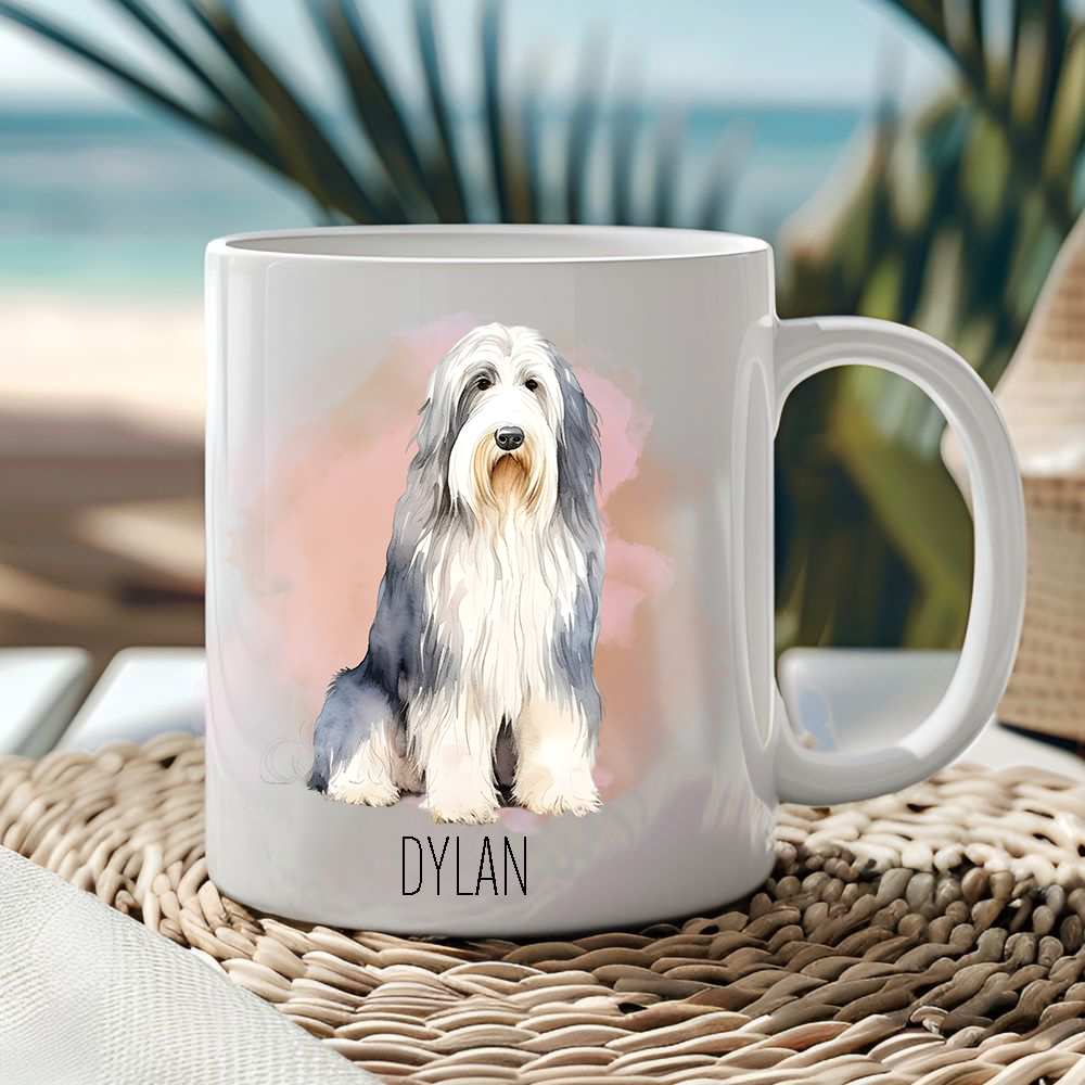 White ceramic mug showing a Bearded Collie in a sitting position with a pink/peachy smoke effect background.  Mug is placed on a wicker mat  and a seascape lurks behind.