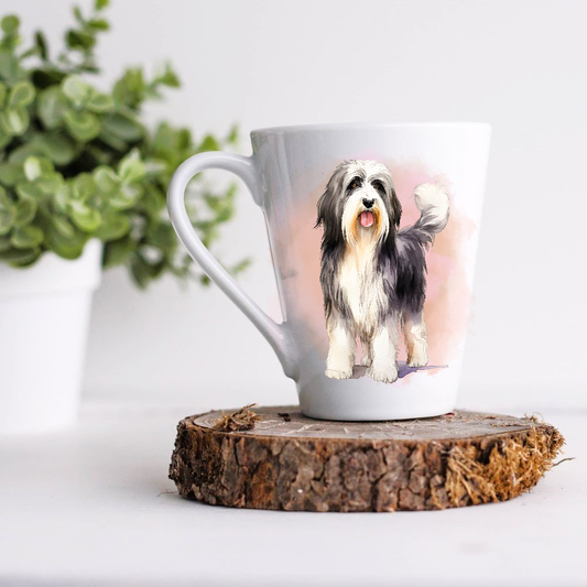 A Bearded Collie is shown standing on a white ceramic latte mug in a pink misty background.  The mug is placed on a wooden disc with a green foliage plant in a white pot to the left hand side.
