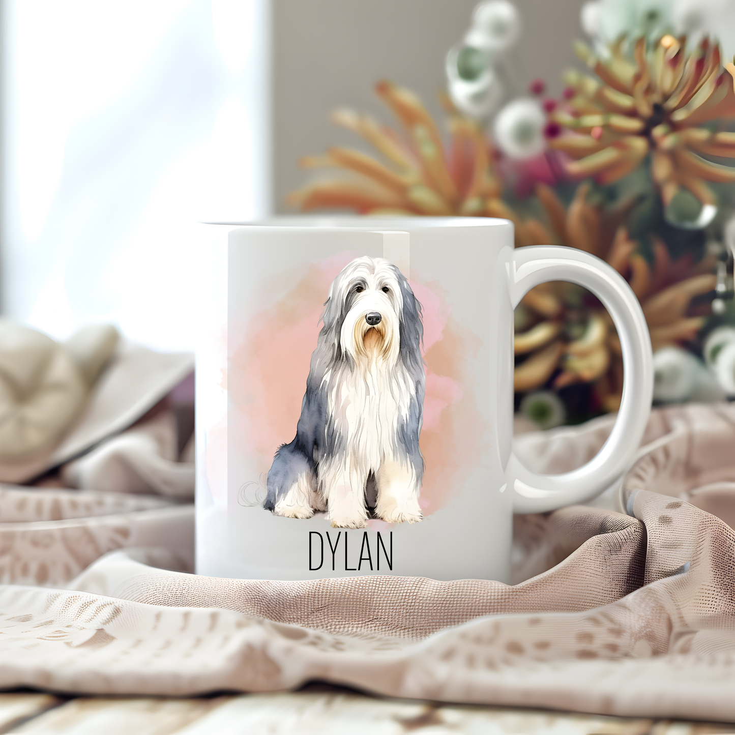 Personalised coffee mug featuring a seated Bearded Collie on a white ceramic mug which has been placed on a beige cloth.  Stems of large  flowers accessorizes the image. 