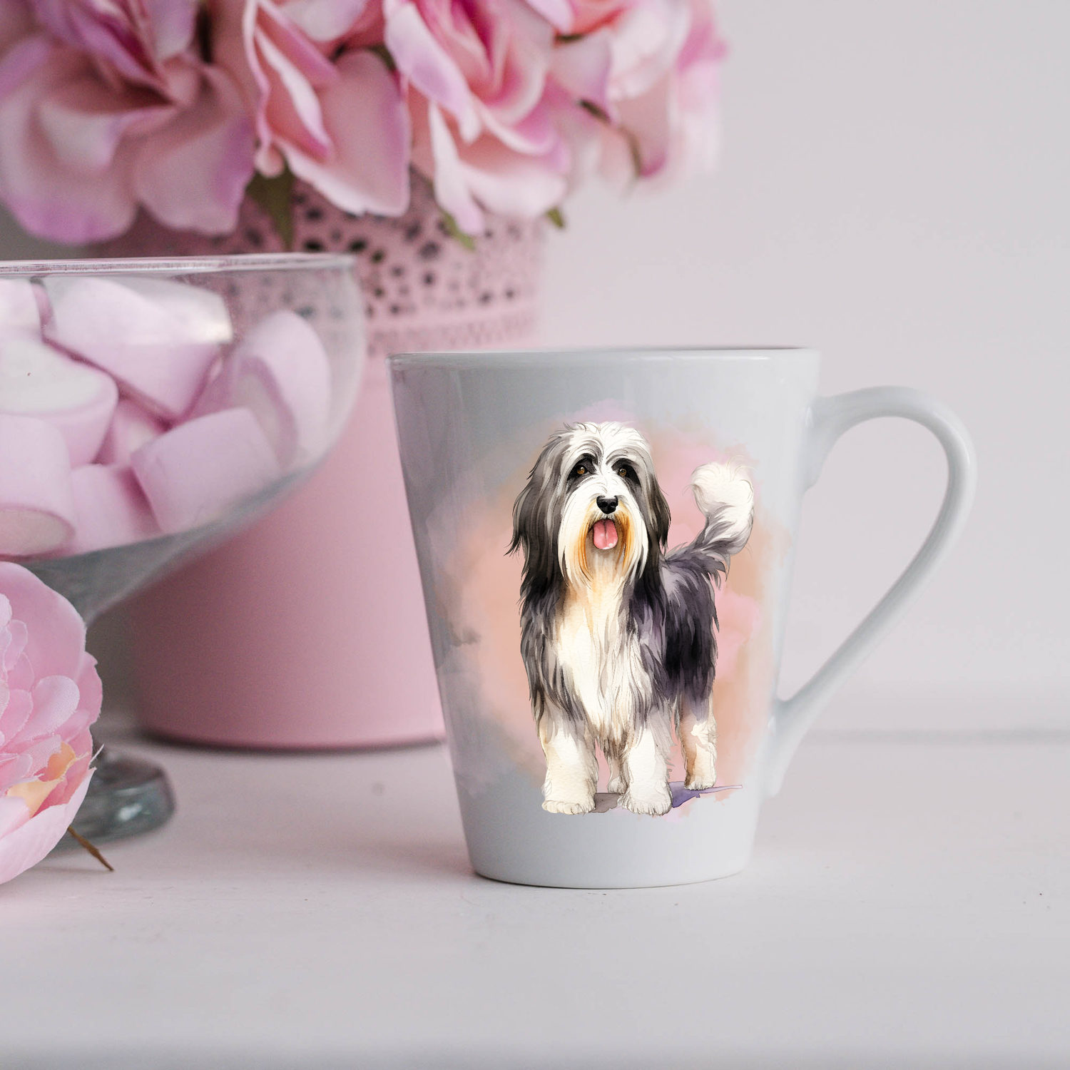 A Bearded Collie is shown lying on a white ceramic latte mug in a smoky background and looking forward.  The mug is placed on a white surface with pink flowers in a pink plant pot and a dish of pink marshmallows.