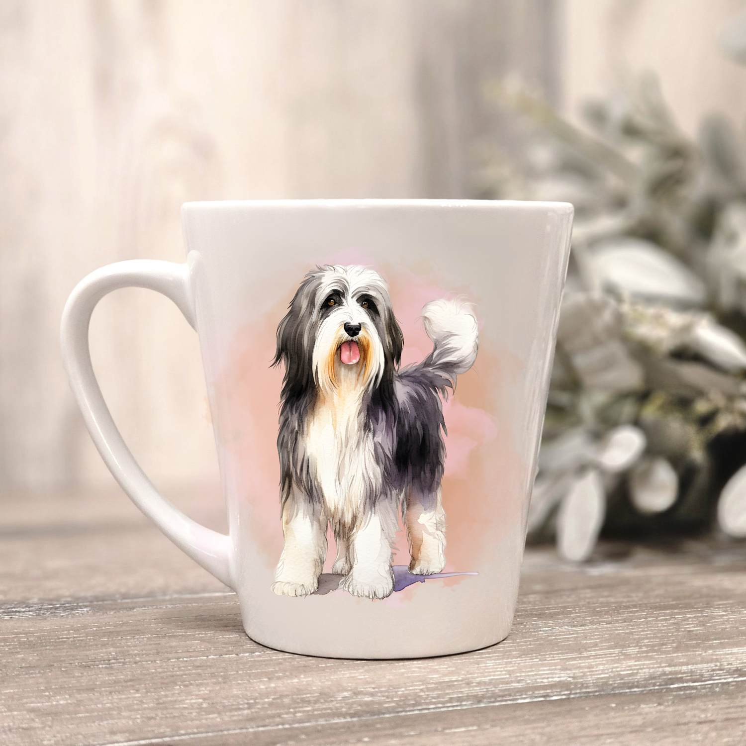 A Bearded Collie is shown lying on a white ceramic latte mug in a smoky background and looking forward.  The mug is placed on a wooden surface with sage green foliage to the right hand side.