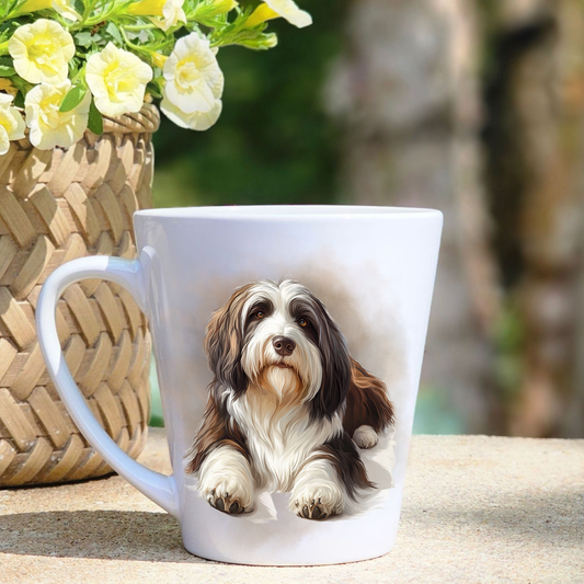 A white ceramic latte mug featuring a  Bearded Collie lying down among a smoky mist background.  A basket containing yellow flowers sits to the left hand side.
