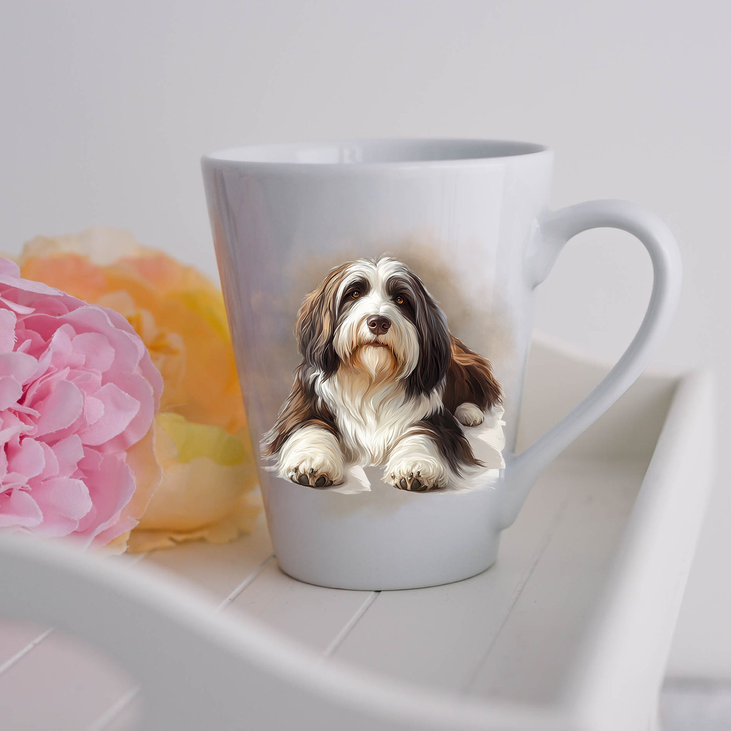 A Bearded Collie is shown lying on a white ceramic latte mug in a smoky background and looking forward.  The mug is placed on a white tray with pink and yellow flowers. 