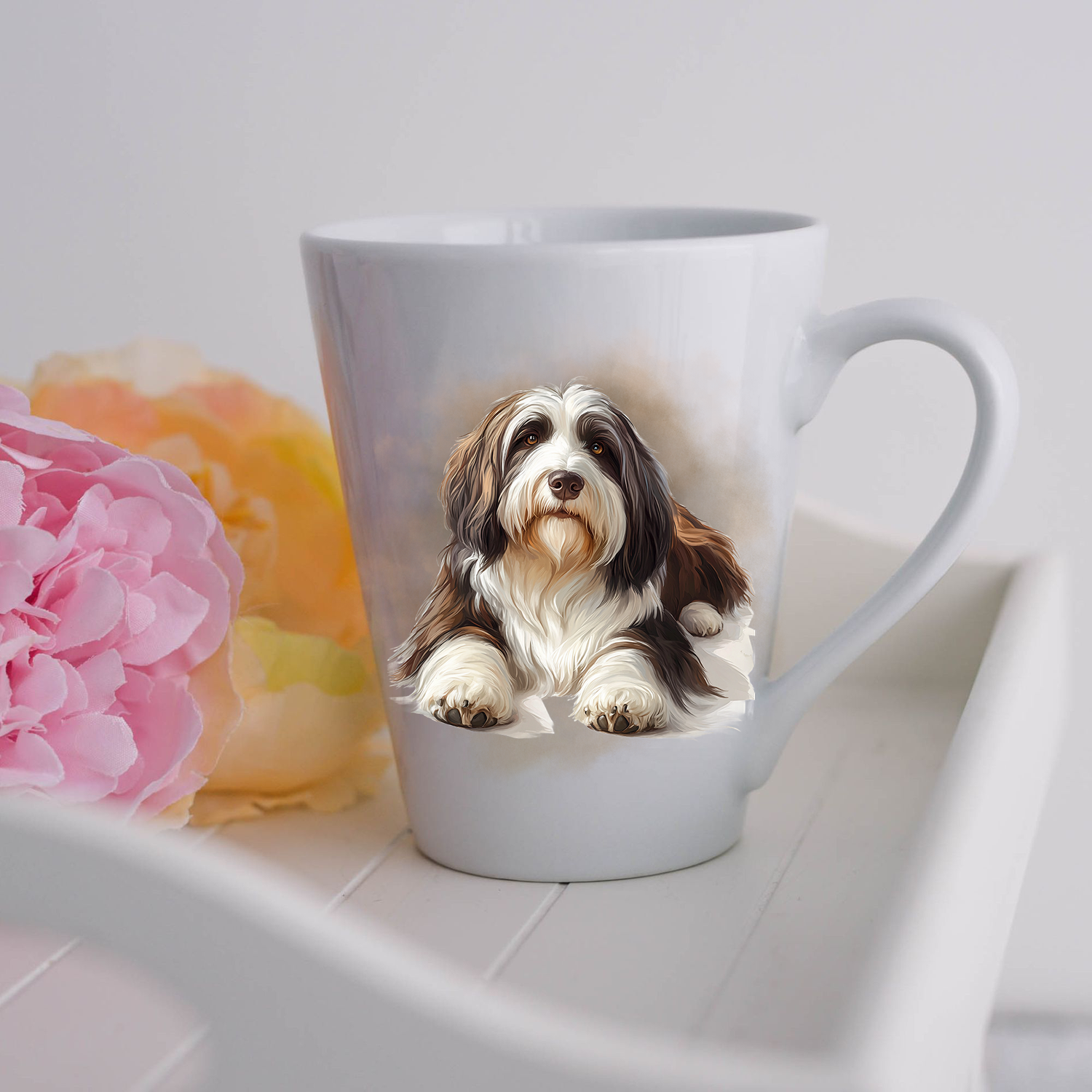 A Bearded Collie is shown lying on a white ceramic latte mug in a smoky background and looking forward.  The mug is placed on a white tray with pink and yellow flowers. 