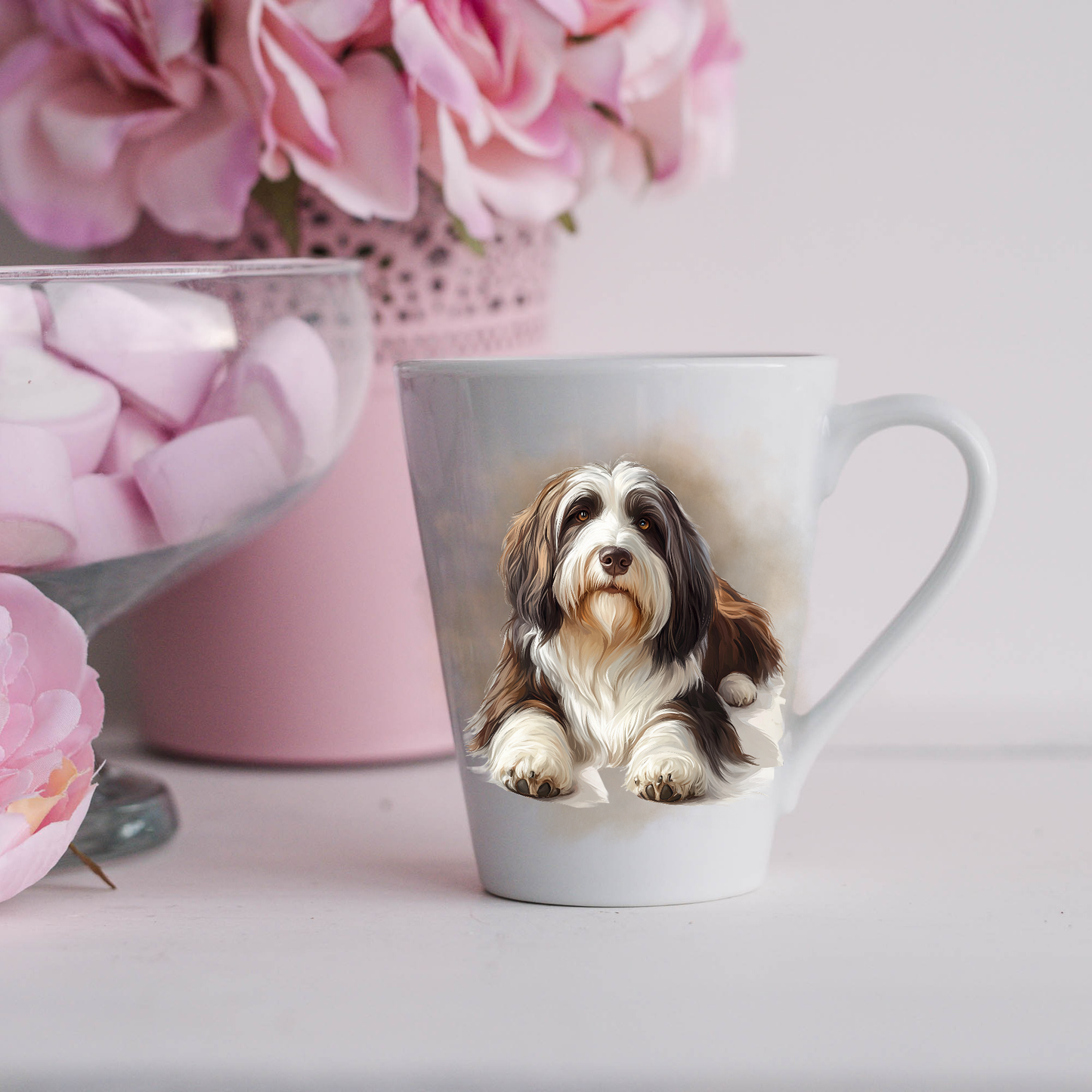 A Bearded Collie is shown lying on a white ceramic latte mug in a smoky background and looking forward.  The mug is placed on a white surface with pink flowers in a pink plant pot and a dish of pink marshmallows. 