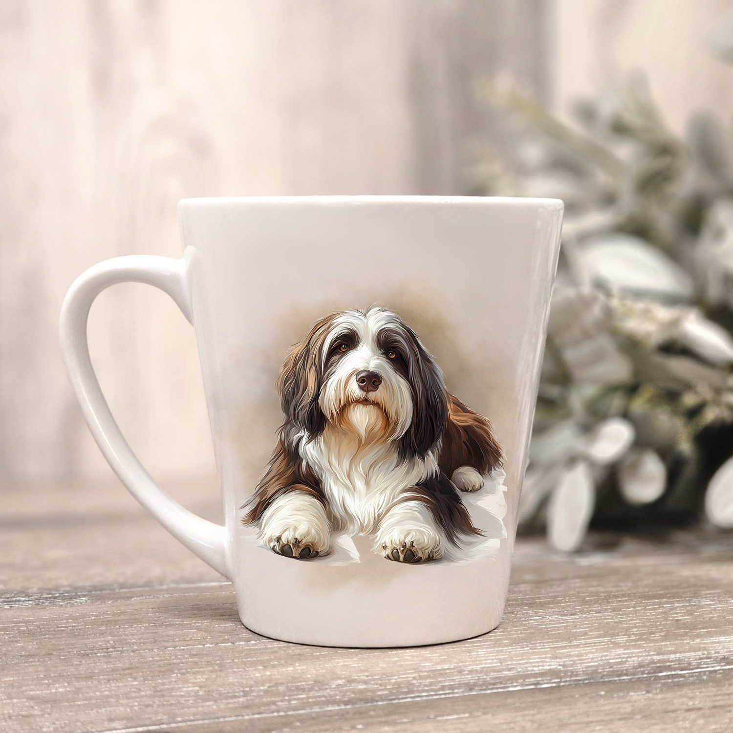 A Bearded Collie is shown lying on a white ceramic latte mug in a smoky background and looking forward.  The mug is placed on a wooden surface with sage green foliage to the right hand side.