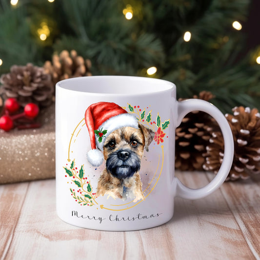 A ceramic mug with a watercolour design of a Border Terrier wearing a Santa hat, positioned on a wooden surface with Christmas decorations around.