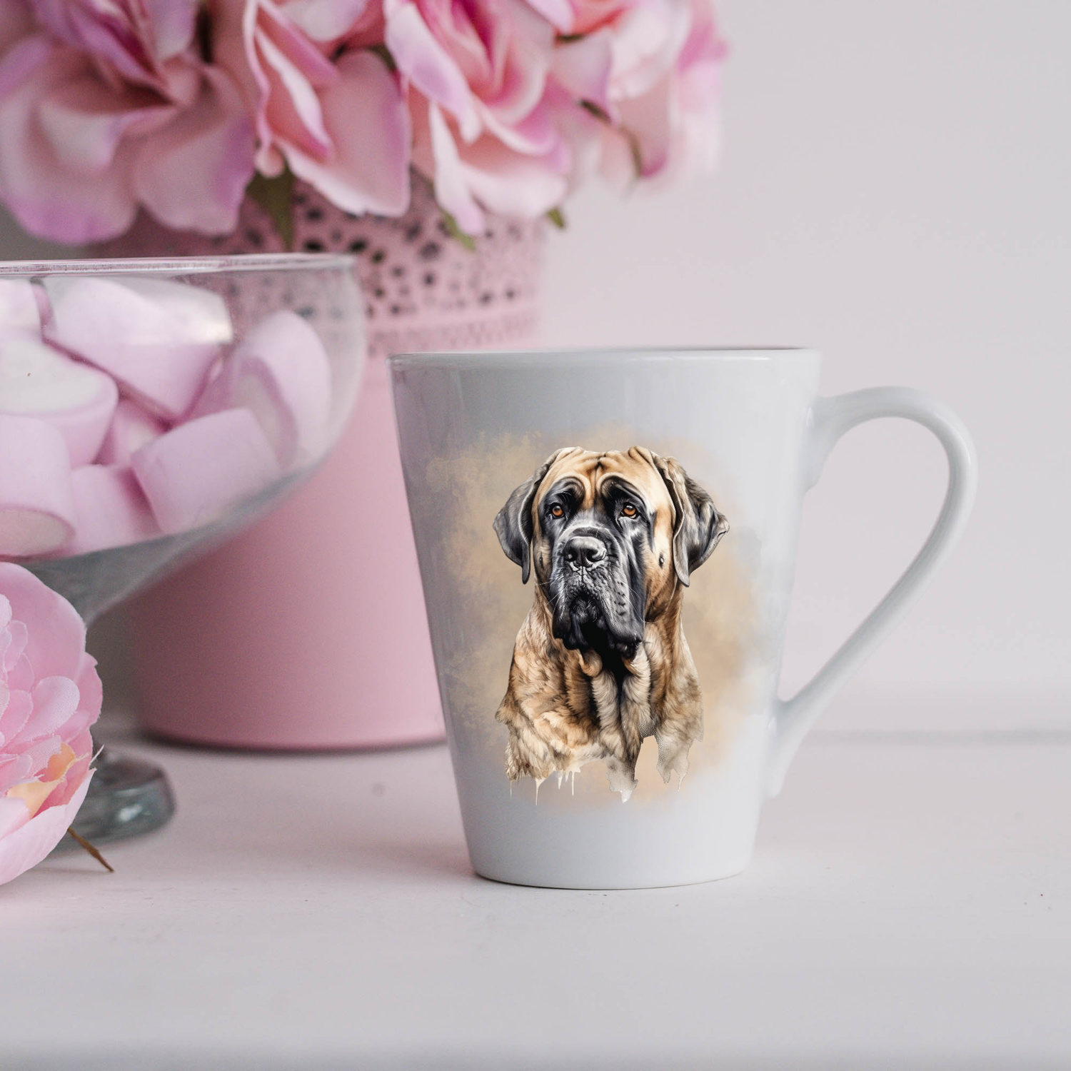 A Bullmastiff is shown on a white ceramic latte mug in a smoky background and looking forward.  The mug is placed on a white surface with pink flowers in a pink plant pot and a dish of pink marshmallows.