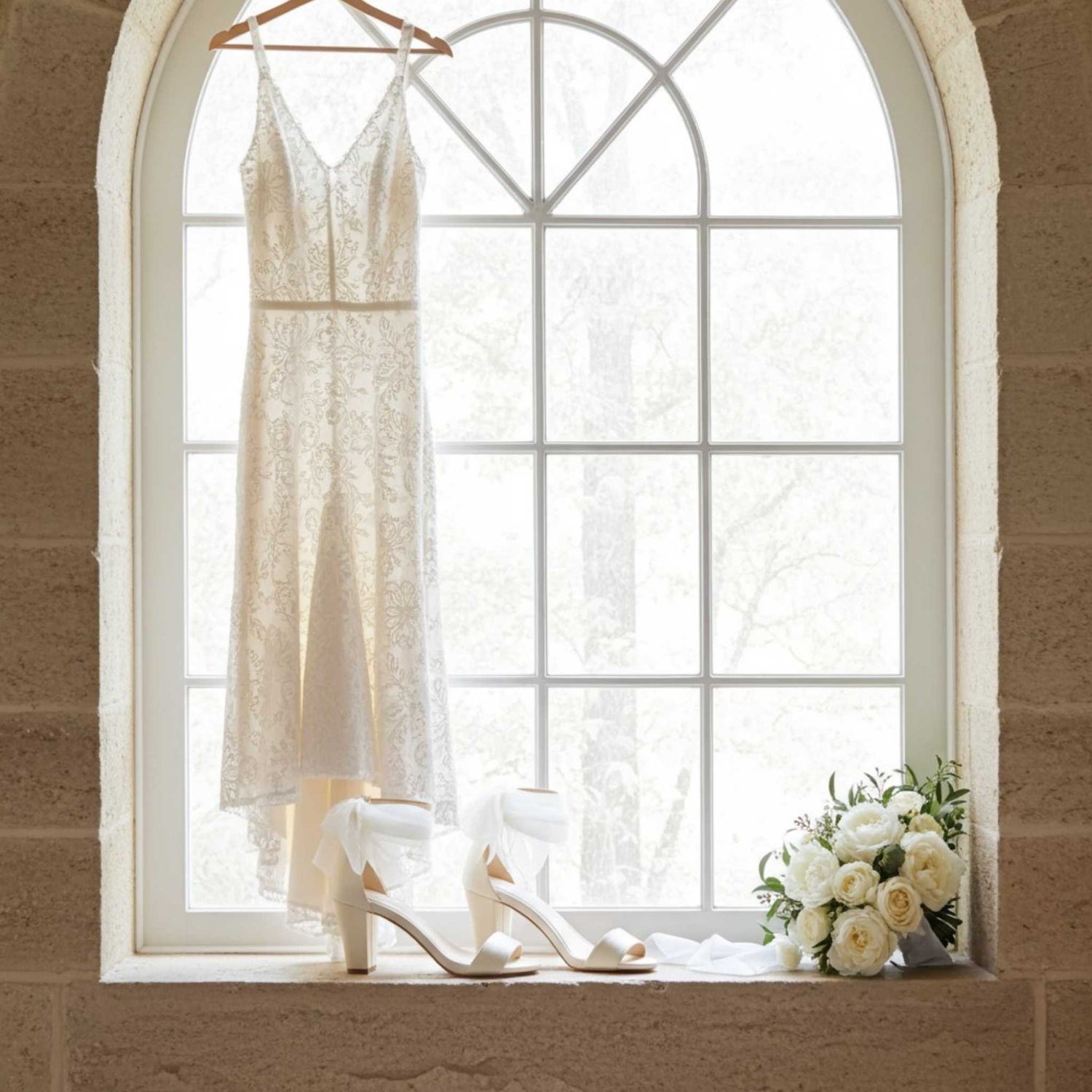 Bridal dress on a hanger with block heel bridal shoes and flowers on a windowsill.