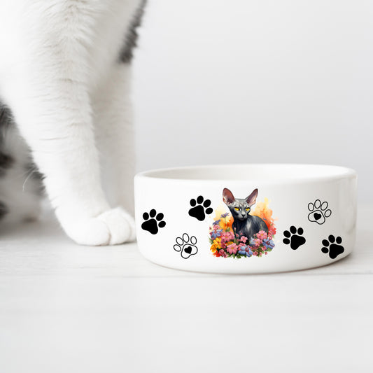 white ceramic cat food or water bowl featuring a black sphynx cat with a floral theme and paw prints. A white cat leg is in the background.