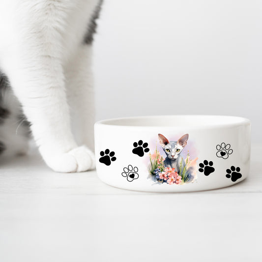 white ceramic cat food or water bowl featuring a platinum sphynx cat with a floral theme and paw prints. A white cat leg is in the background.