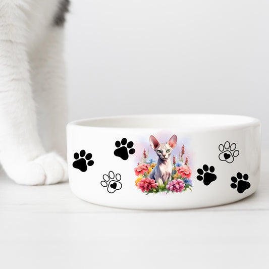 white ceramic cat food or water bowl featuring a cream sphynx cat with a floral theme and paw prints. A white cat leg is in the background.
