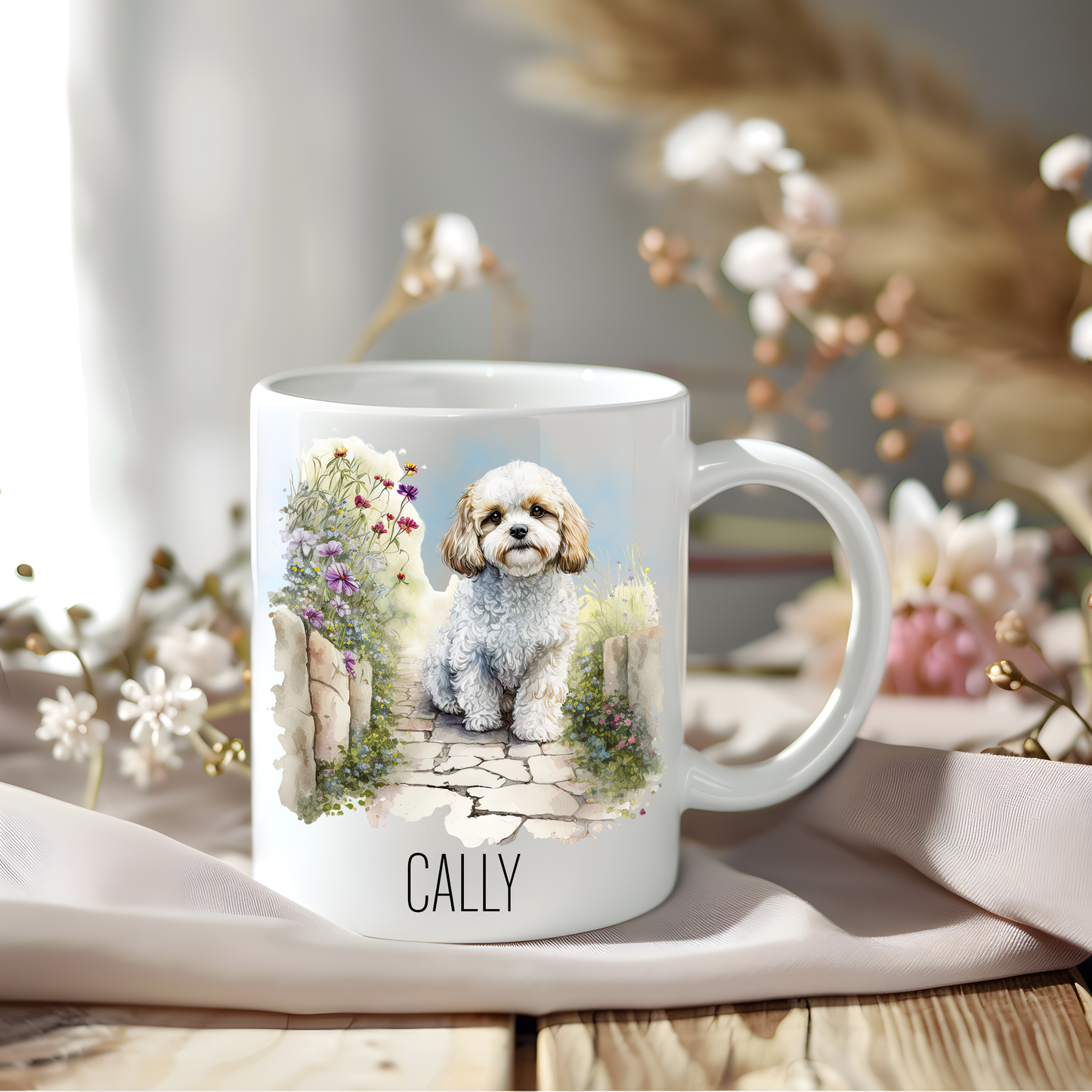 Cavachon coffee mug personalised with a pet name.  The mug is sat on a wooden surface with a creamy coloured cloth and various flowers adorn the background.