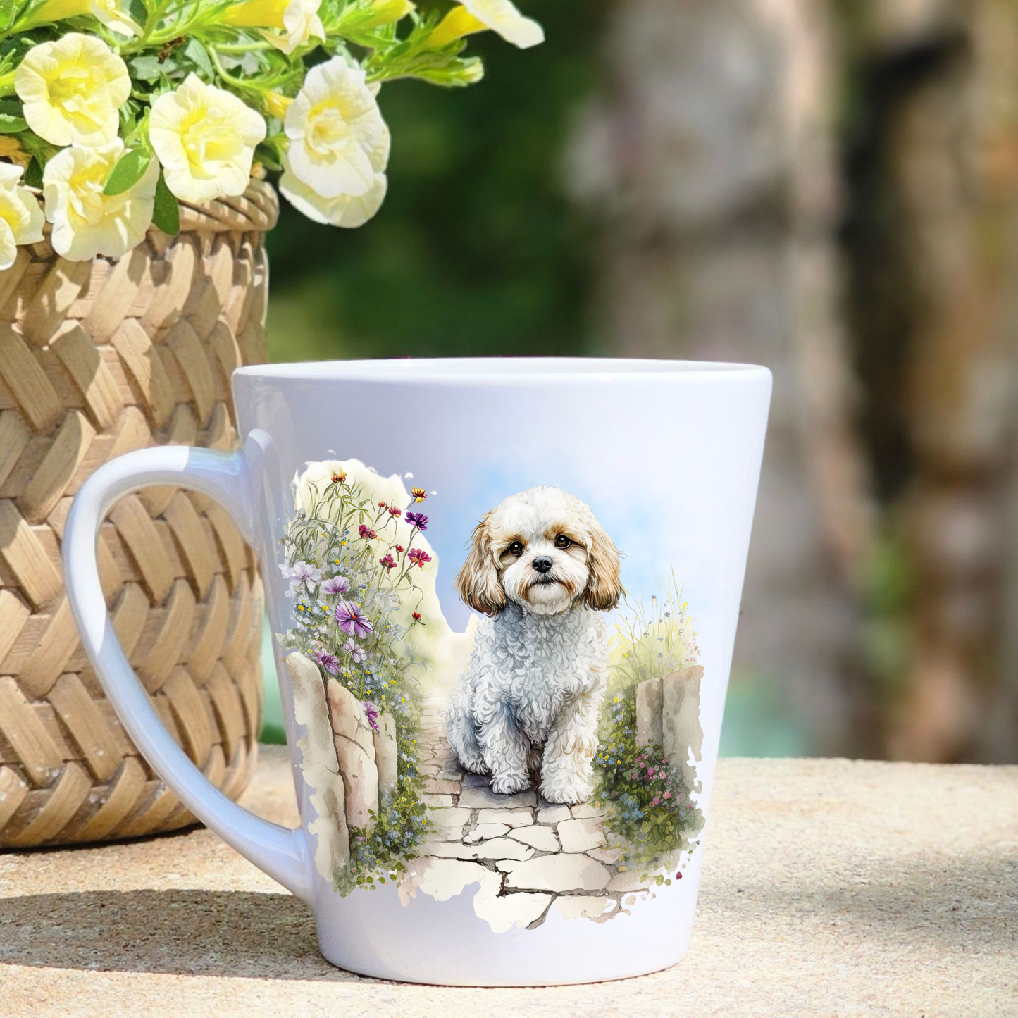 A white ceramic latte mug featuring a seated Cavachon in a garden setting.  A basket containing yellow flowers sits to the left-hand side.