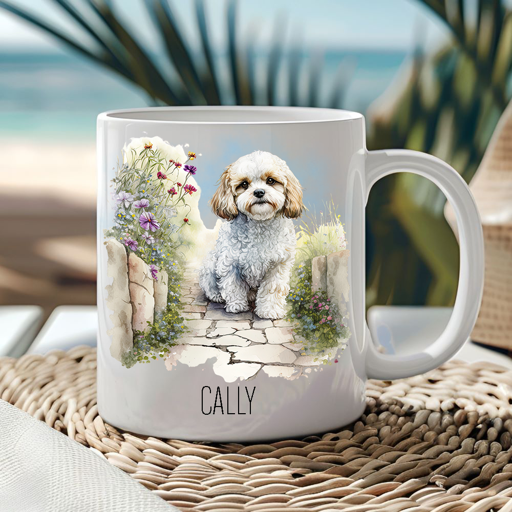 White ceramic mug showing a Cavachon sitting in a garden with the Mug placed on a wicker mat and a seascape sits behind.
