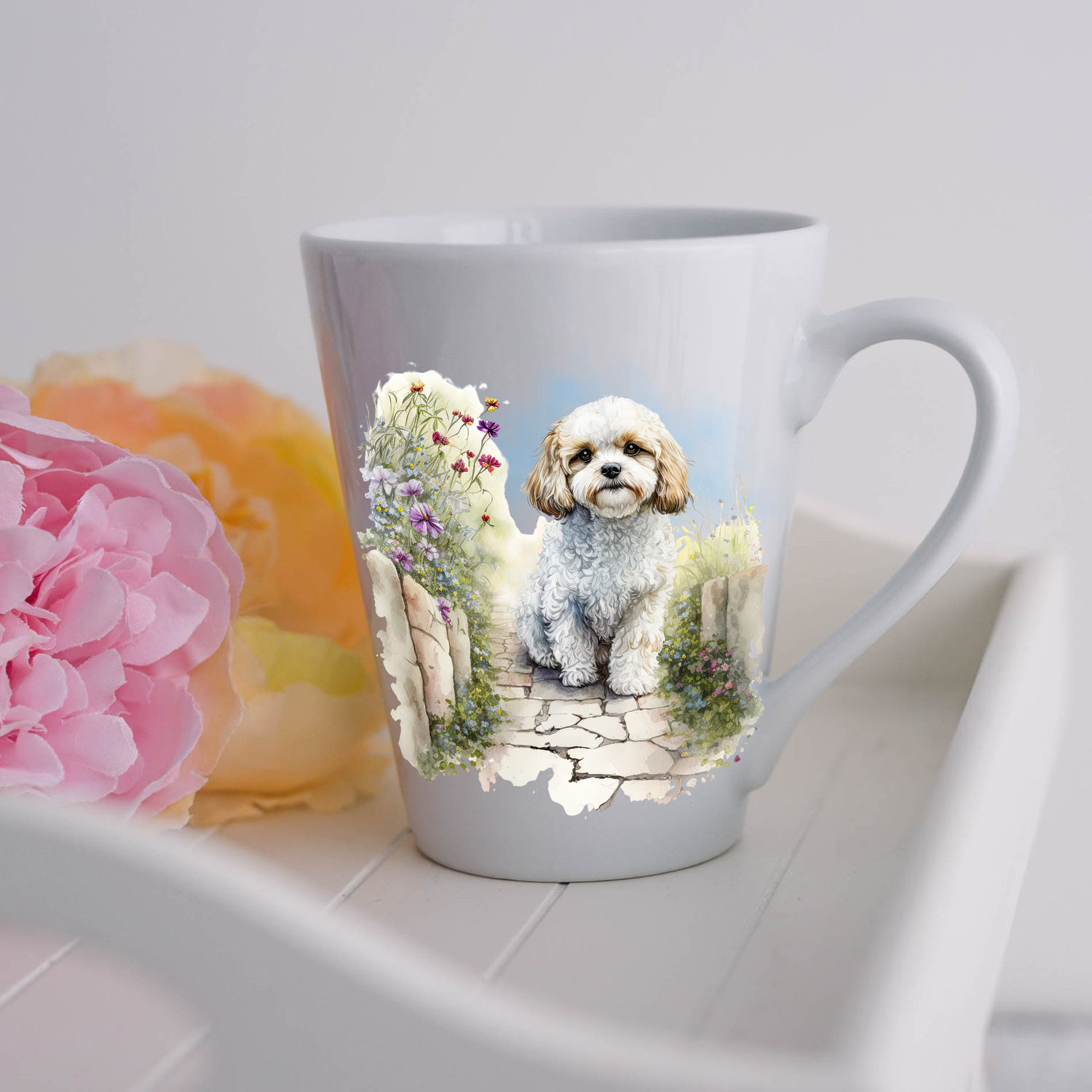 A Cavachon is shown sitting in a garden on a white ceramic latte mug and looking forward.  The mug is placed on a white tray with pink and yellow flowers. 