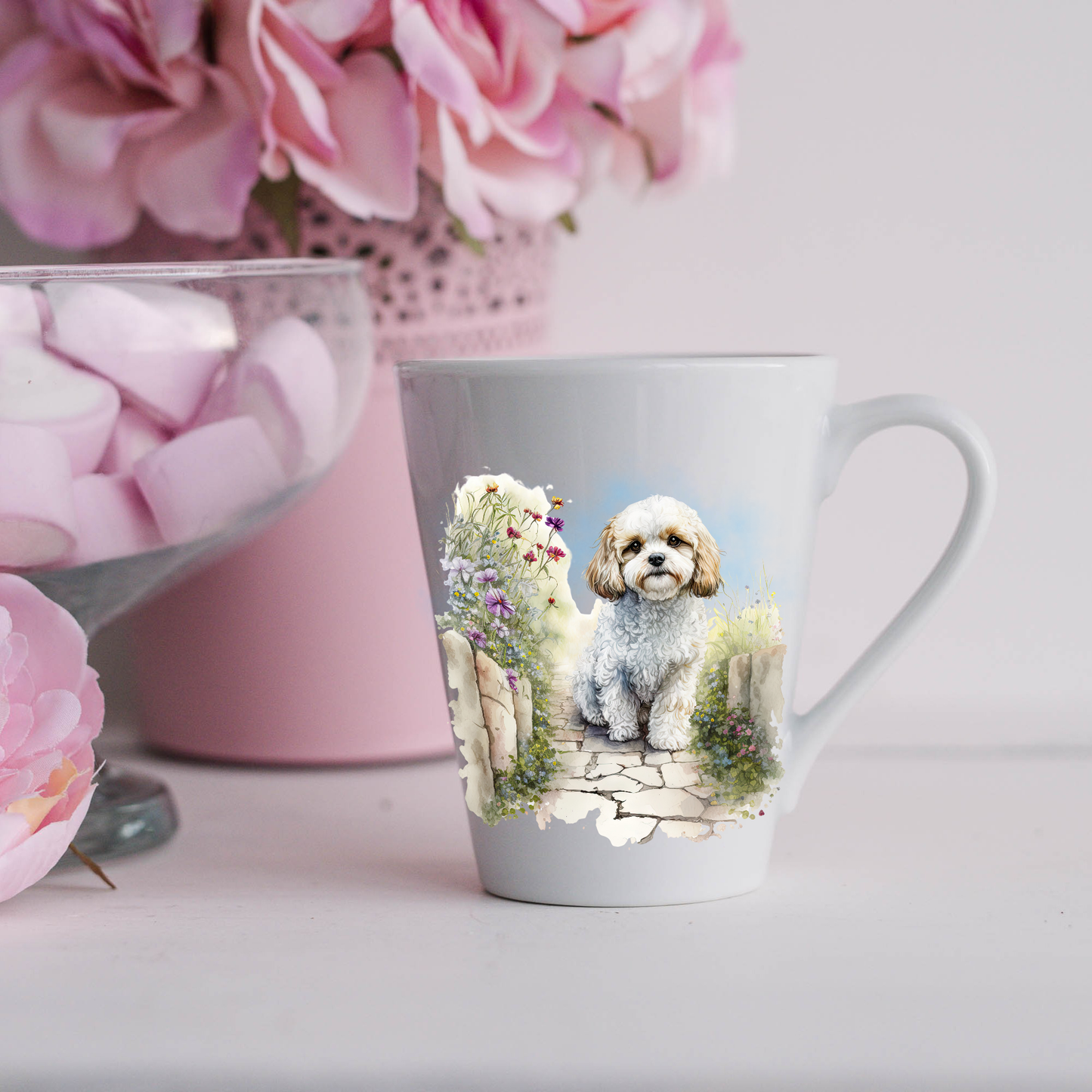 A Cavachon dog is shown sitting on a paved pathway in a garden on a white ceramic latte mug.  The mug is placed on a white surface with pink flowers in a pink plant pot and a dish of pink marshmallows.