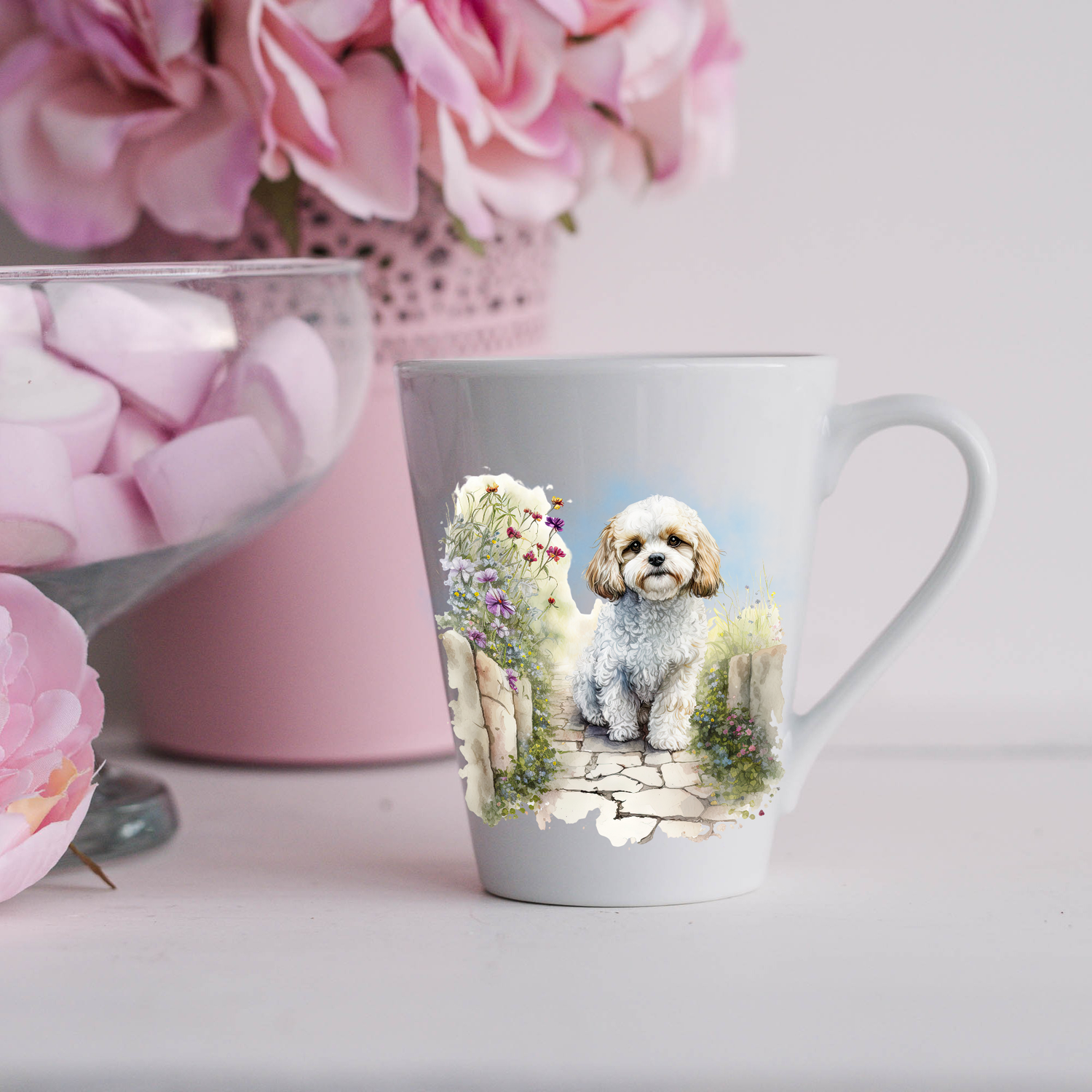 A Cavachon dog is shown sitting on a paved pathway in a garden on a white ceramic latte mug.  The mug is placed on a white surface with pink flowers in a pink plant pot and a dish of pink marshmallows.