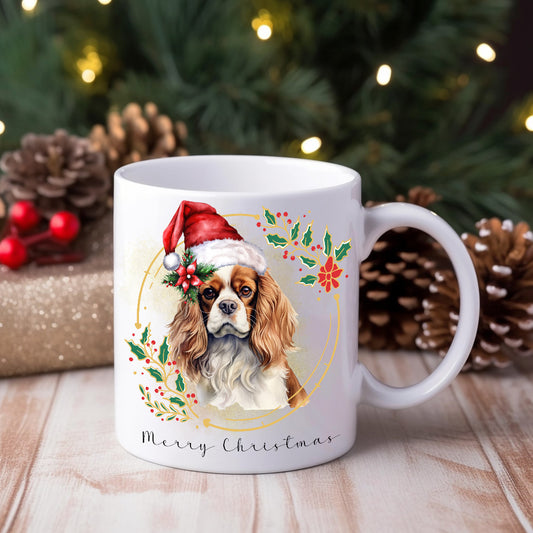 White ceramic mug featuring a Cavalier King Charles Spaniel wearing a Santa hat on a Christmas themed mug positioned on a wooden surface with a Christmas tree in the background.