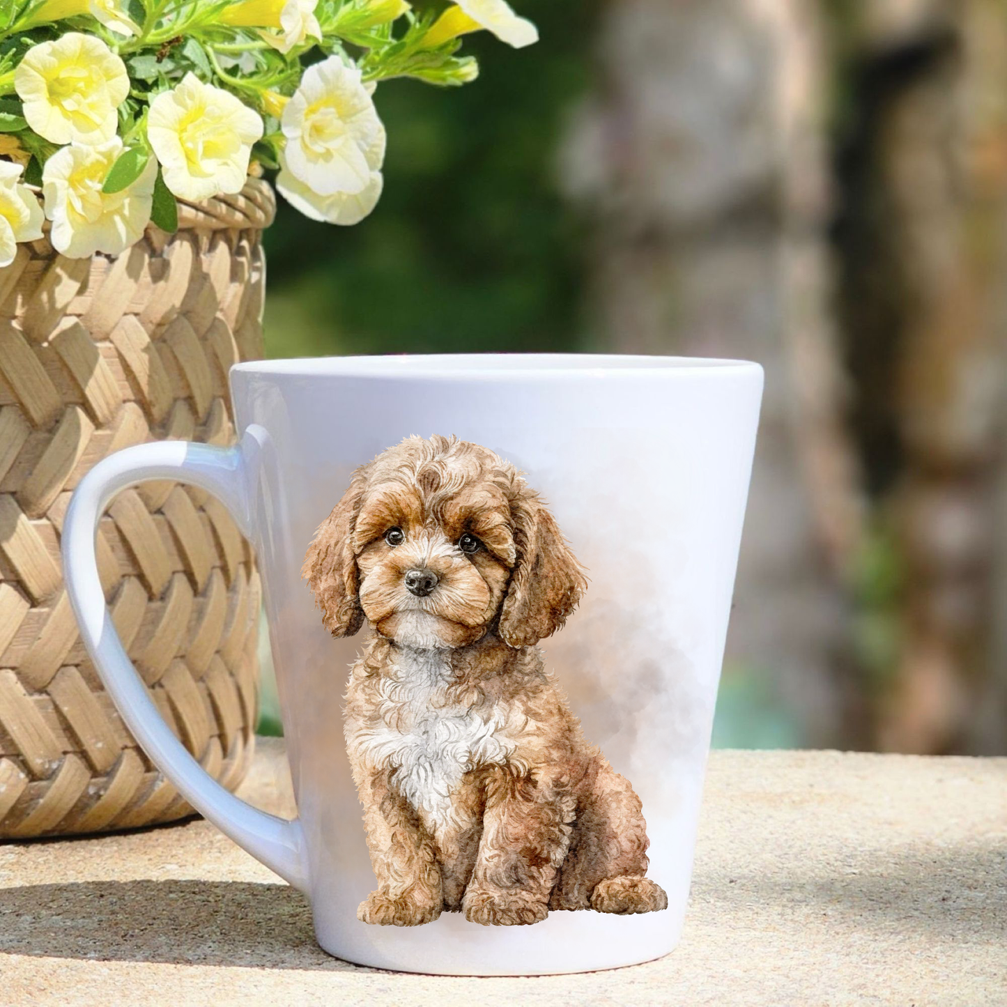 A white ceramic latte mug featuring a seated Cavapoo in a beige misty background.  A basket containing yellow flowers sits to the left-hand side.