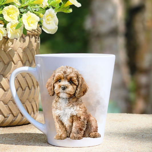 A white ceramic latte mug featuring a seated Cavapoo in a beige misty background. A basket containing yellow flowers sits to the left-hand side.