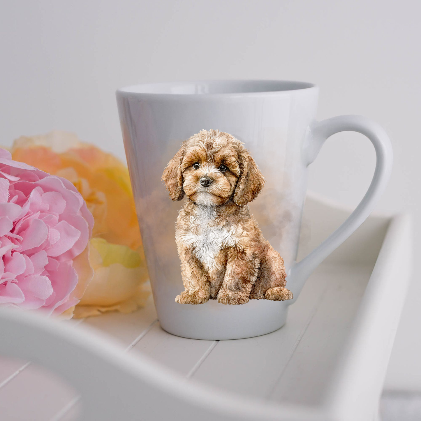 A Cavapoo dog is shown sitting on a white ceramic latte mug in a smoky background and looking forward.  The mug is placed on a white tray with pink and yellow flowers. 