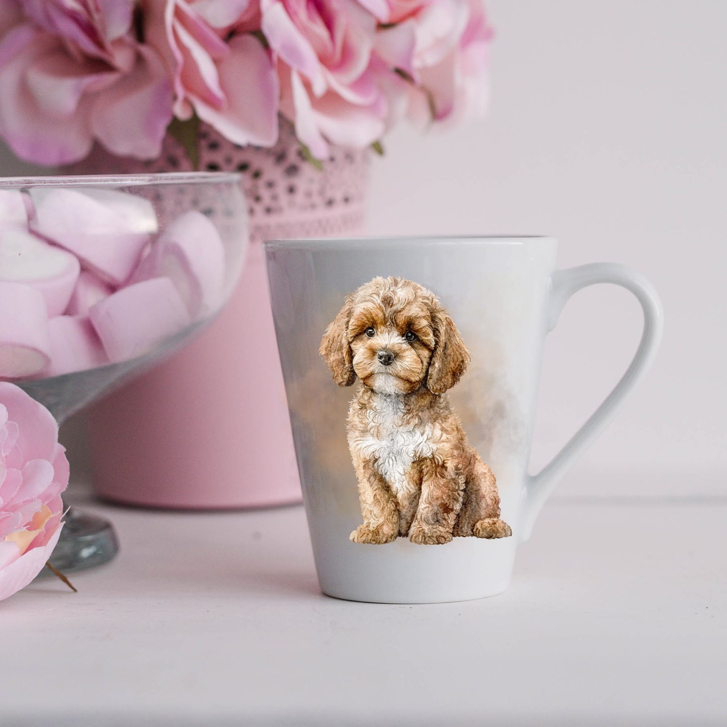 A Cavapoo is shown sitting on a white ceramic latte mug in a smoky background and looking forward.  The mug is placed on a white surface with pink flowers in a pink plant pot and a dish of pink marshmallows.
