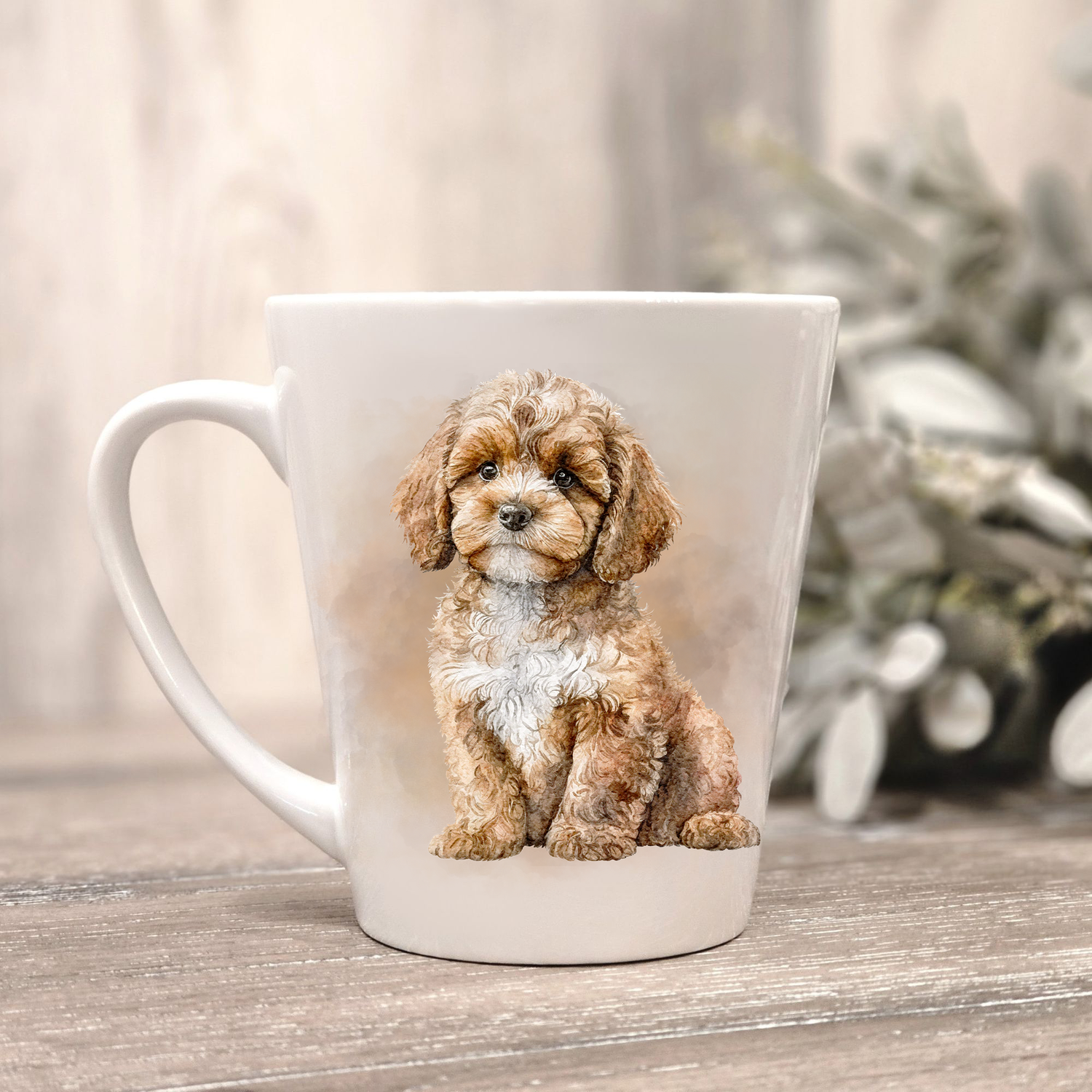 A Cavapoo dog is seen in a seated position on a white ceramic latte mug in a smoky background and looking forward.  The mug is placed on a wooden surface with sage green foliage to the right-hand side.