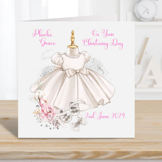 Personalised Christening card for a girl. Christening gown with bow and shoes with a floral arrangement. Card placed on a wooden surface with Alphabet letters and a clock in the background.