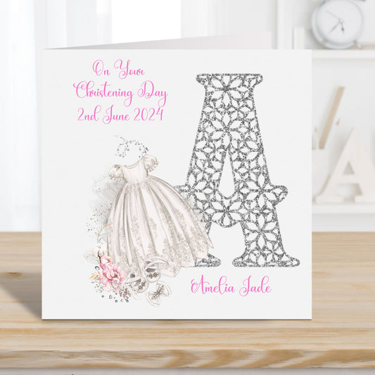 Personalised Christening card for a Girl featuring a large initial and a Christening gown, shoes and pink flowers.