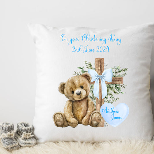 Personalised White Christening Cushion for a boy featuring a teddy bear sitting beside a cross with a blue ribbon bow and green foliage.