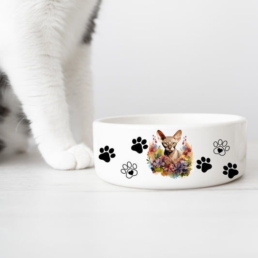 white ceramic cat food or water bowl featuring a cinnamon sphynx cat with a floral theme and paw prints. A white cat leg is in the background.