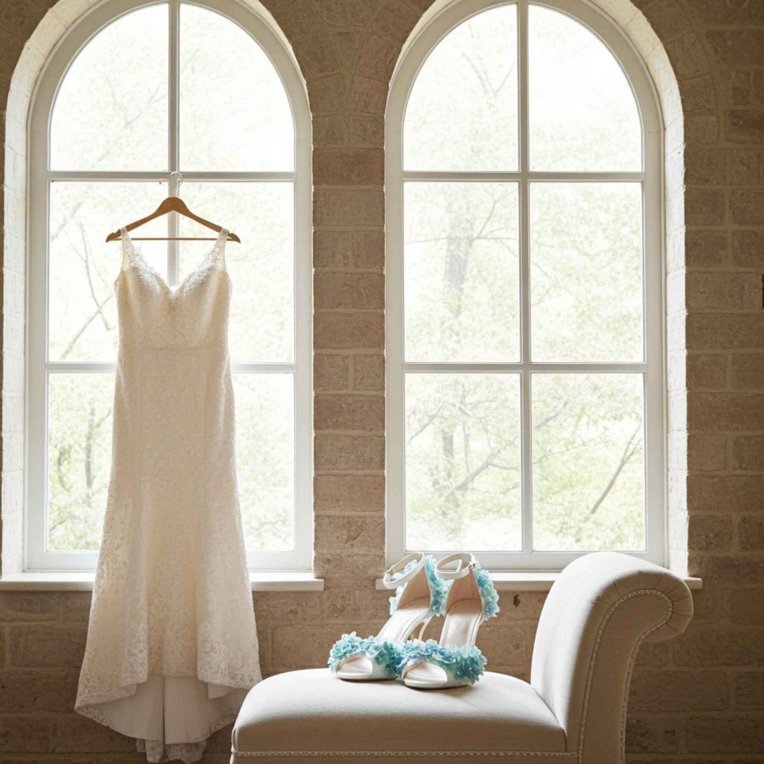 White wedding dress hanging on a hanger next to a white chair with blue floral bridal sandals on a brick wall background.