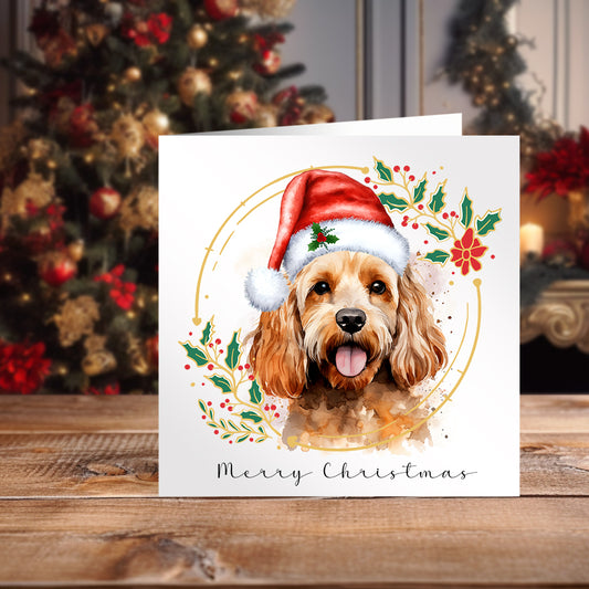 Apricot Cockapoo wearing a Santa hat on a Christmas themed card positioned on a wooden surface in front of a Christmas Tree.