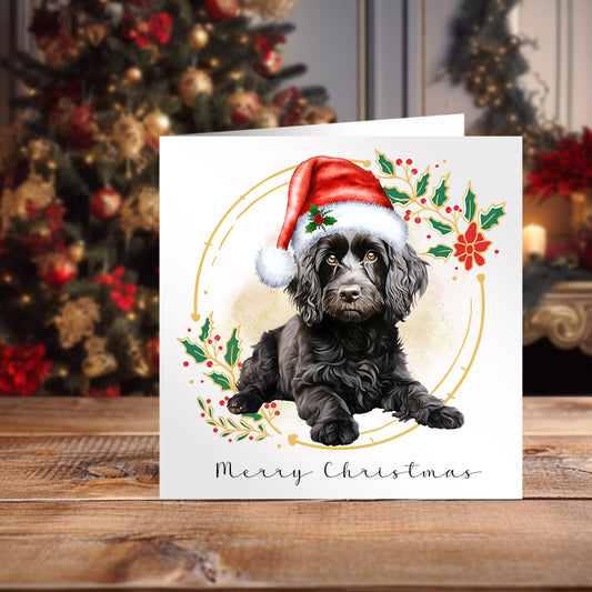 Black Cockapoo wearing a Santa hat lying down on a Christmas themed card on a wooden surface in front of a decorated Christmas tree.