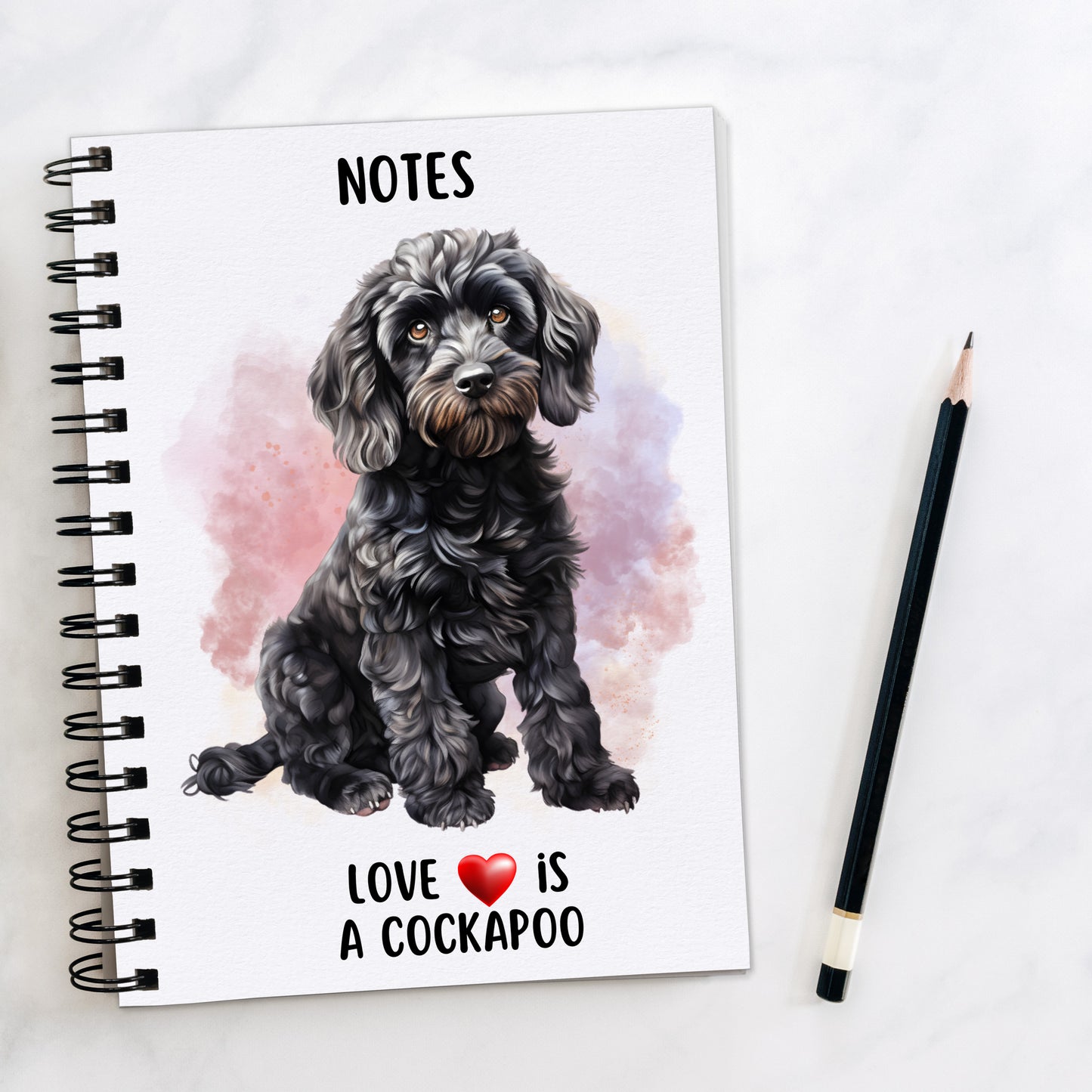 A5 white notebook with black spiral wire binding.  A black Cockapoo sits on the front.  A smoky pink and lilac background decorates and a black pencil is placed to the right hand side.  A quote with a red heart is placed at the bottom of the book.