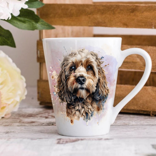 A watercolour headshot of a Cockapoo dog facing forward on a white latte mug. The mug is positioned on a wooden surface and in front of a brown crate. A cream flower is to the left hand side.