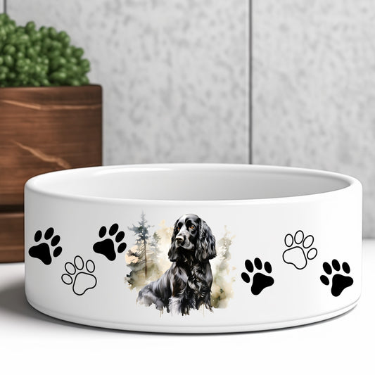 white ceramic dog food or water bowl featuring a black cocker spaniel dog and paw prints. A brown wooden box and green plant are in the background.
