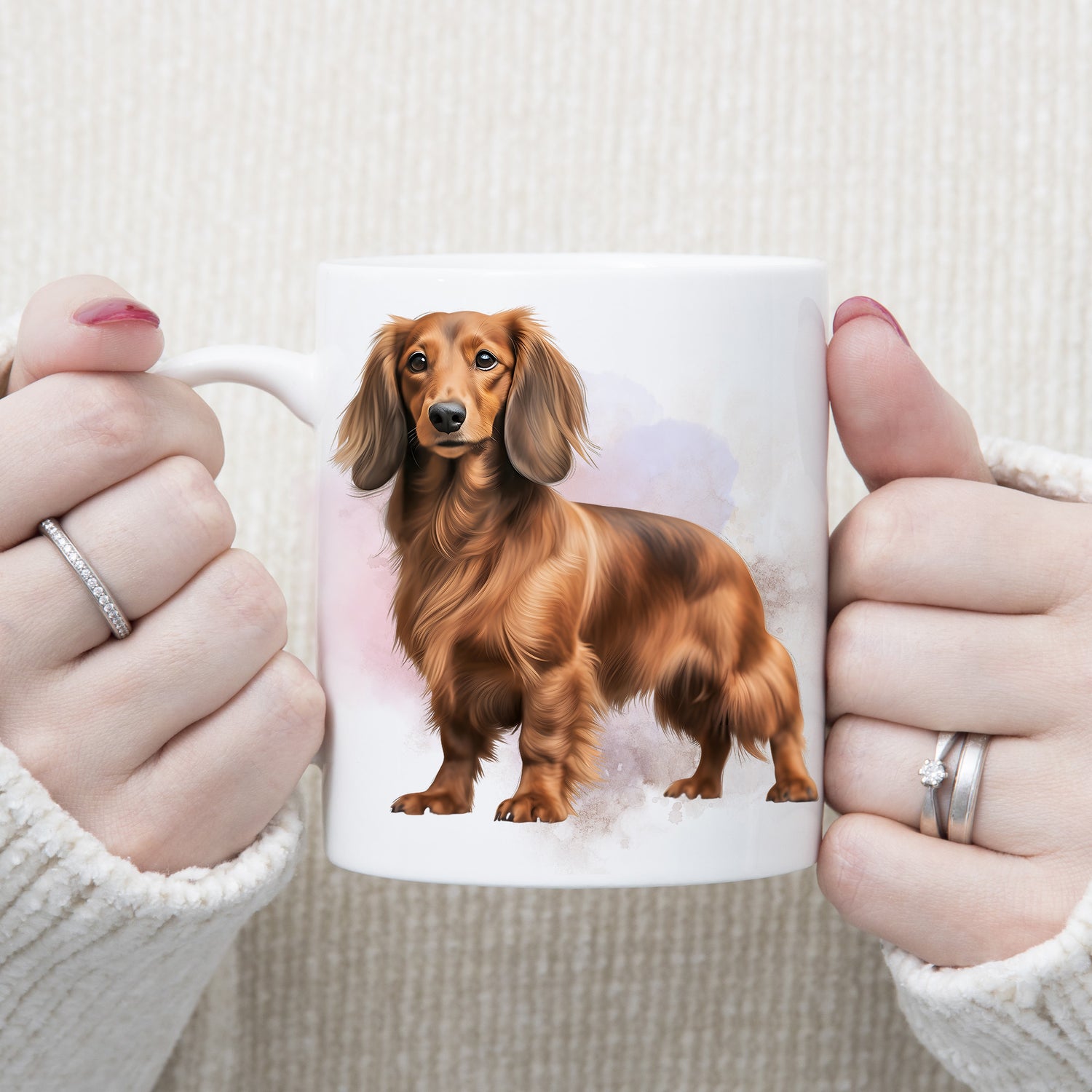 A long haired Dachshund is standing against a lilac and pink background on this white ceramic mug.  The mug is being held by a woman with both hands.