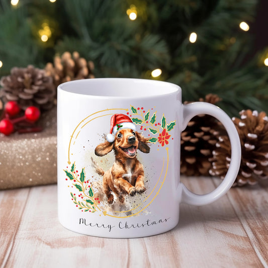 A white ceramic mug with a watercolour design of a Dachshund wearing a Santa hat, positioned on a wooden surface with Christmas decorations around, including pine cones and lights.