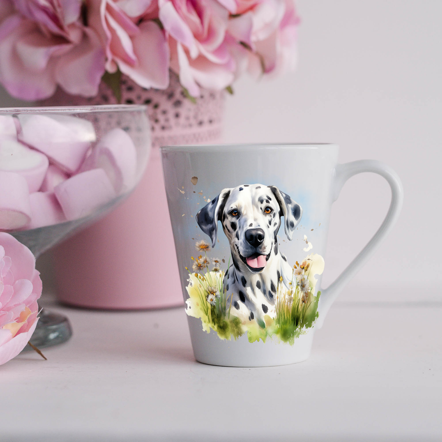 A Dalmatian is shown on a white ceramic latte mug in grass and wildflower scene.  The mug is placed on a white surface with pink flowers in a pink plant pot and a dish of pink marshmallows.
