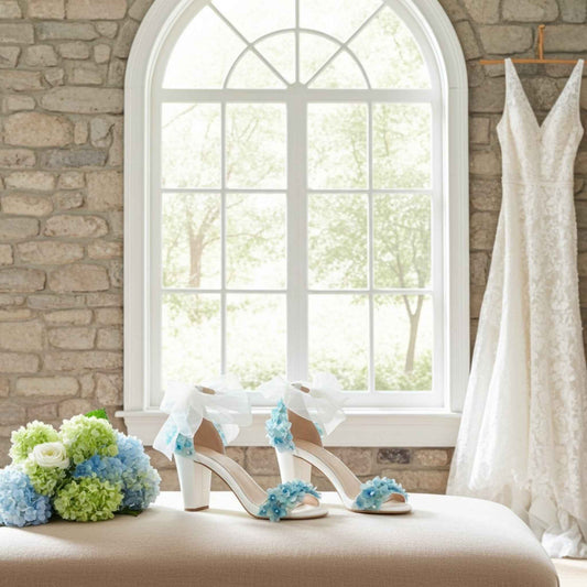 Open toe block heel floral bridal shoes with a lace wedding gown, blue flowers on a cushion in front of a stone wall and window.