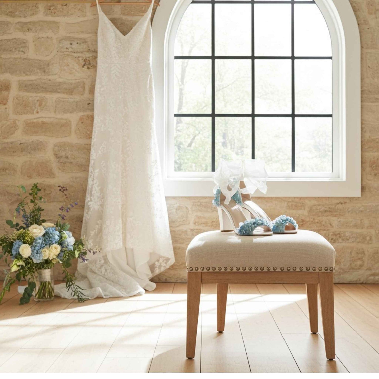 Ivory wedding dress hanging on a rail with floral high-heeled bridal shoes and a bouquet of flowers on a stool in a room with a large window.