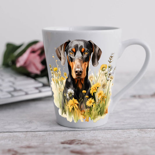 A 12oz white ceramic latte mug with a printed image of a black and tan Doberman facing forward and sitting among yellow flowers. A computer keyboard sits in the background.  The mug is positioned on a wooden surface.