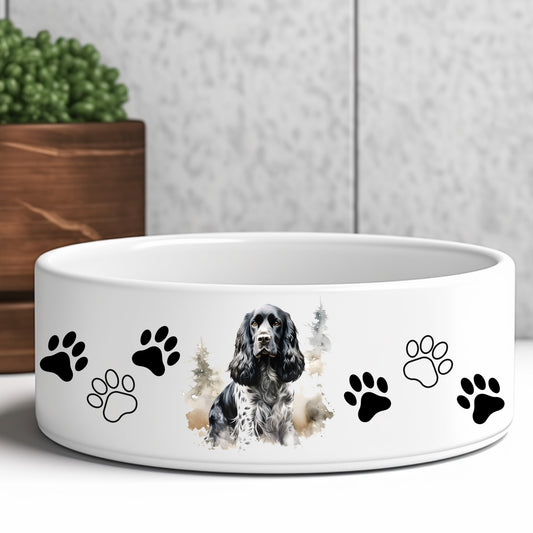 white ceramic dog food or water bowl featuring a black and white parti-colour cocker spaniel dog and paw prints. A brown wooden box and green plant are in the background.