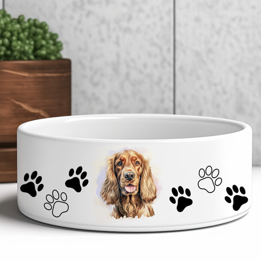white ceramic dog food or water bowl featuring a golden cocker spaniel dog and paw prints. A brown wooden box and green plant are in the background.