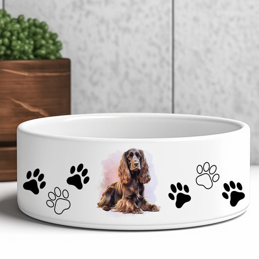 white ceramic dog food or water bowl featuring a liver cocker spaniel dog and paw prints. A brown wooden box and green plant are in the background.