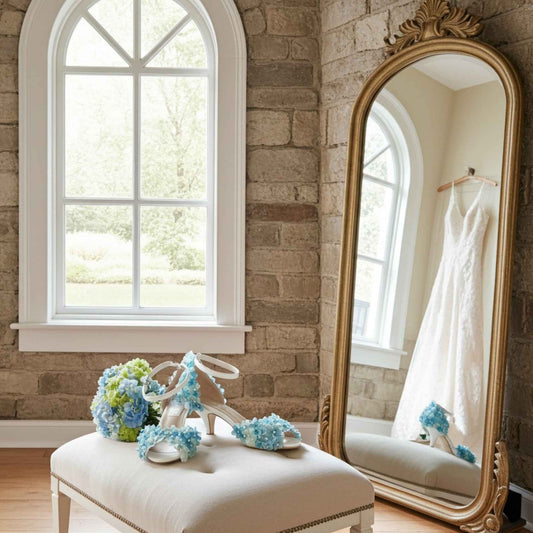 Bridal shoes with floral decorations on a stool next to a large mirror reflecting a wedding dress.