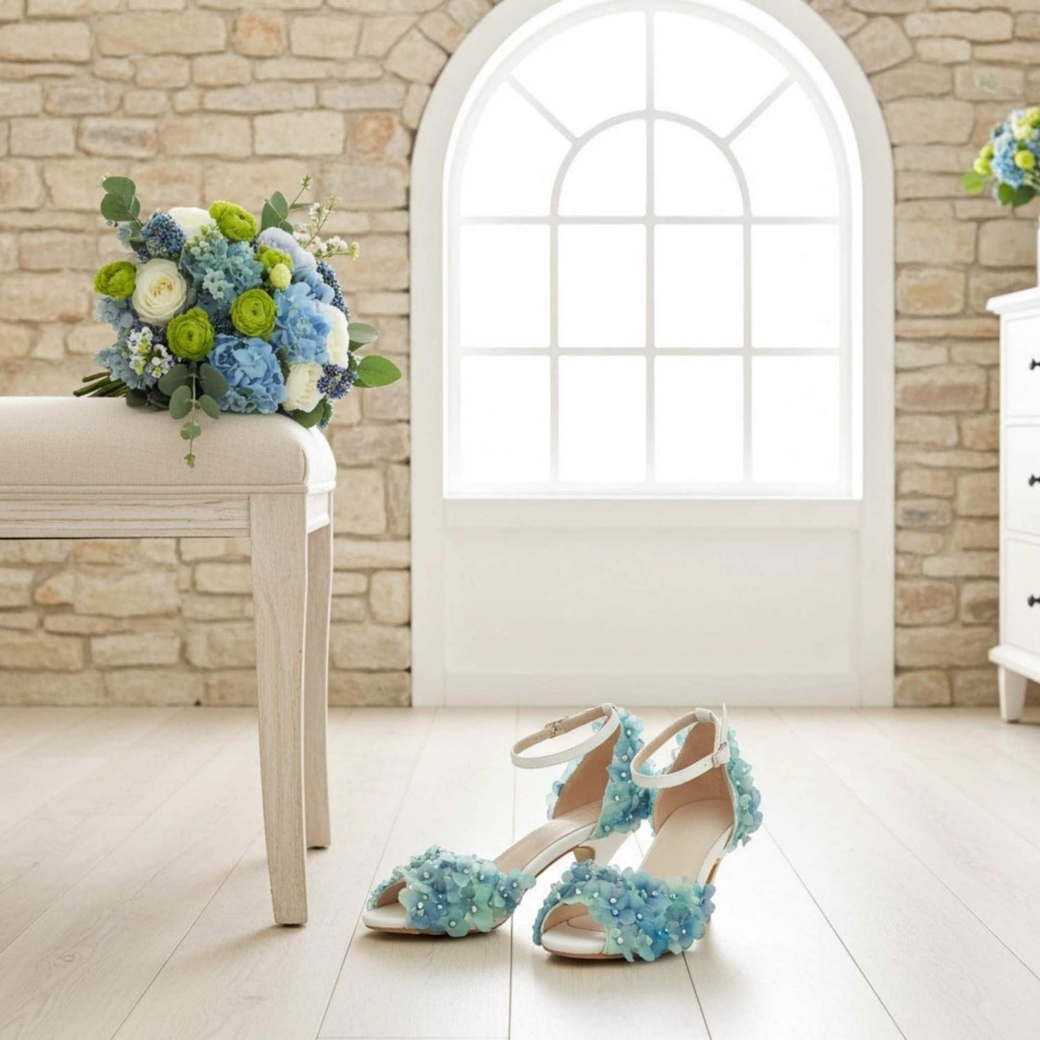 Pair of blue floral bridal sandals on a light wooden floor with a bouquet of flowers in the background.